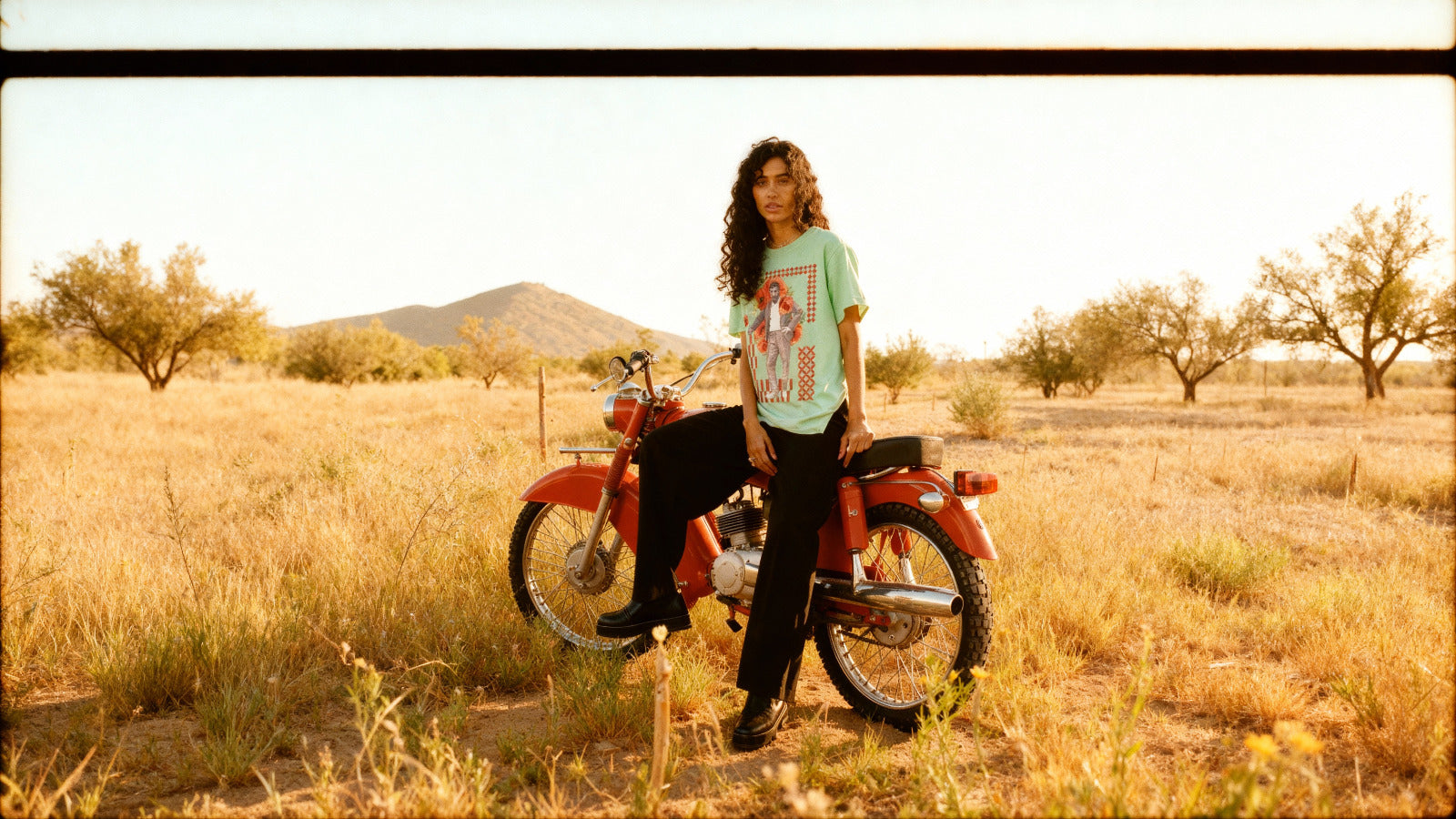 Woman standing next to a red motorcycle in a desert landscape. Arab Hyphenated. 